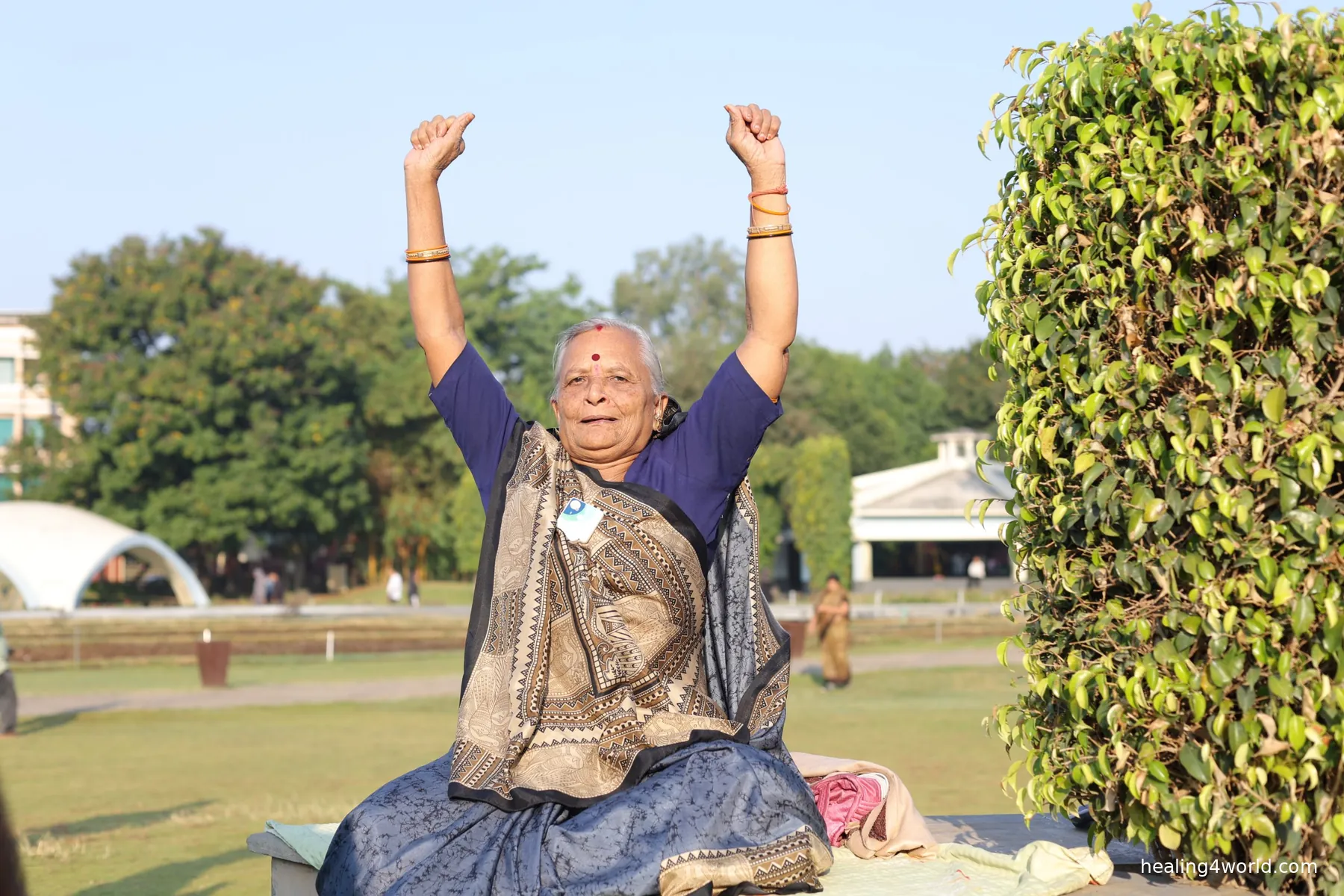 An elder woman in a saree raises both arms in a park, wearing Healy
